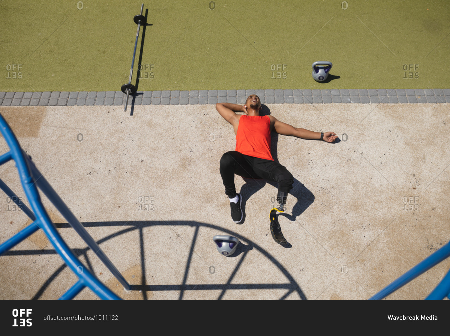 High angle view of disabled mixed race man with a prosthetic running blade working out at outdoor gym wearing wireless earphones, taking a rest lying on his back. Fitness disability healthy lifestyle.
