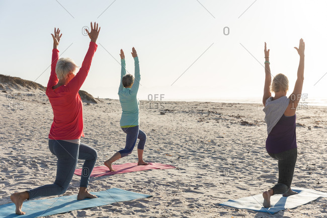 Group of Caucasian female friends enjoying exercising on a beach on a sunny day, practicing yoga and standing in yoga position.