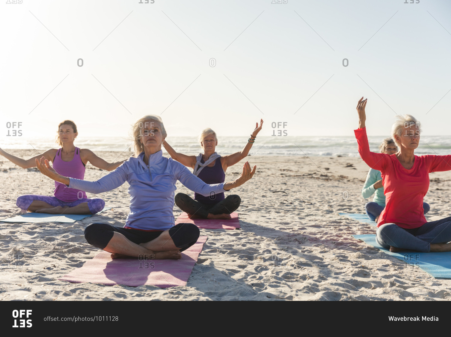 Group of Caucasian female friends enjoying exercising on a beach on a sunny day, practicing yoga, meditating in lotus position with sea in the background.