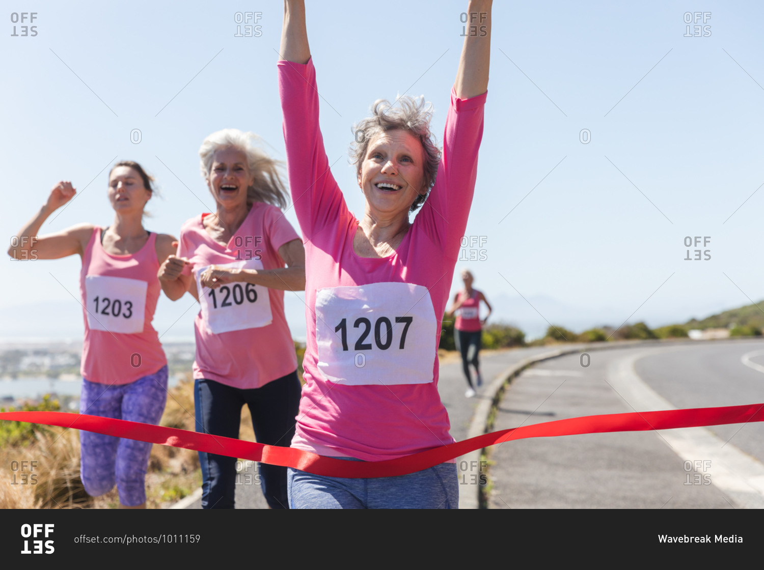 Group of Caucasian female friends enjoying exercising on a sunny day, having running race and wearing numbers, running towards a finish line and celebrating.