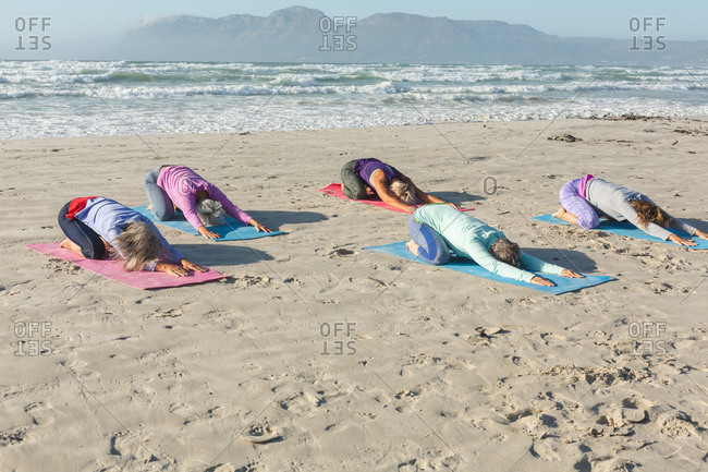 Group of Caucasian female friends enjoying exercising on a beach on a sunny day, practicing yoga and sitting in yoga position.