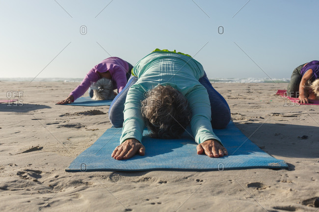 Group of Caucasian female friends enjoying exercising on a beach on a sunny day, practicing yoga and sitting in yoga position.