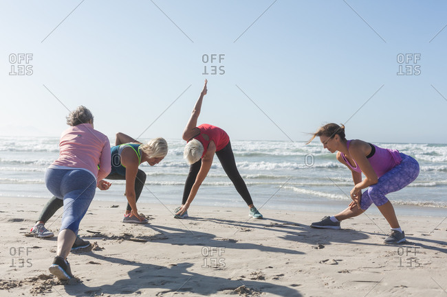 Group of Caucasian female friends enjoying exercising on a beach on a sunny day, practicing yoga and stretching with sea in the background.