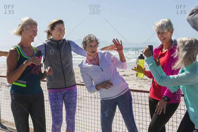 Group of Caucasian female friends enjoying exercising on a beach on a sunny day, taking a break, standing on a promenade and taking photo with a smartphone.