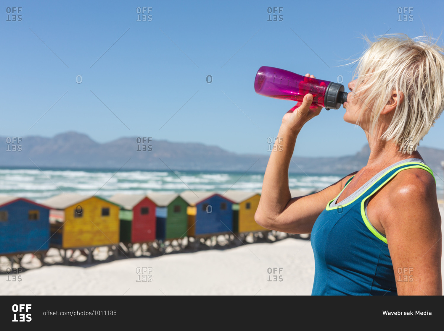 Senior Caucasian woman enjoying exercising on a beach on a sunny day, resting after running on the seashore and drinking water from a bottle with little colorful houses in the background.
