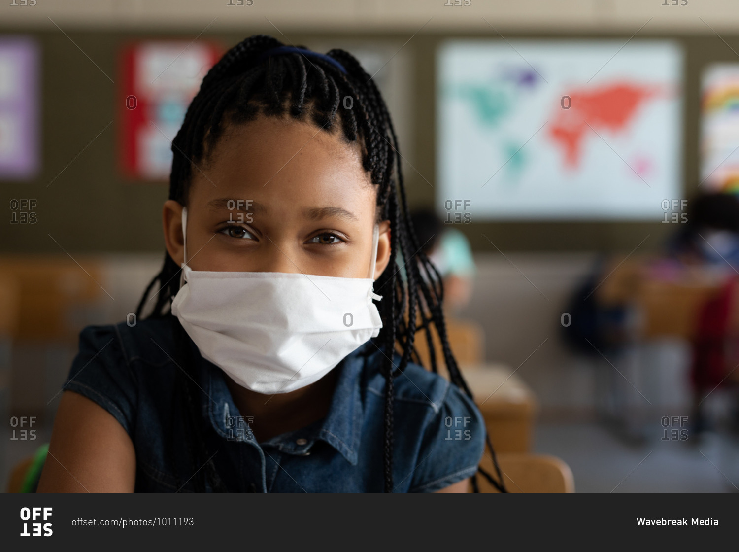 Portrait of a mixed race girl sitting at desks wearing face mask in classroom, looking at camera. Primary education social distancing health safety during Covid19 Coronavirus pandemic.