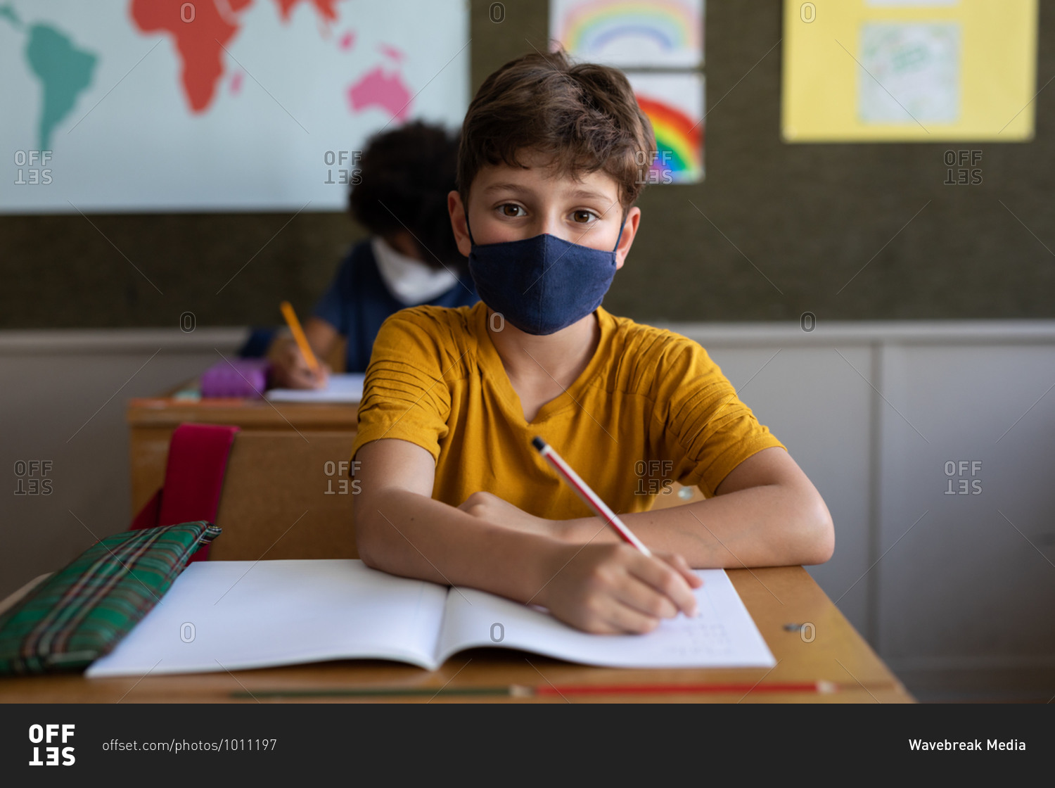 Two multi ethnic children sitting at desks wearing face masks in classroom. Primary education social distancing health safety during Covid19 Coronavirus pandemic.