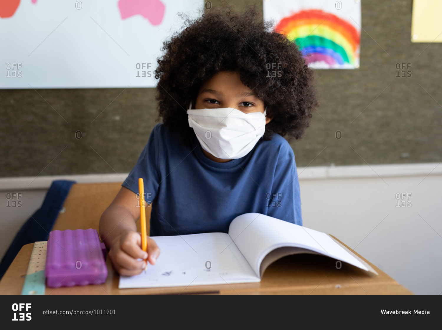 Portrait of a mixed race boy sitting at desk wearing face mask in classroom. Primary education social distancing health safety during Covid19 Coronavirus pandemic.