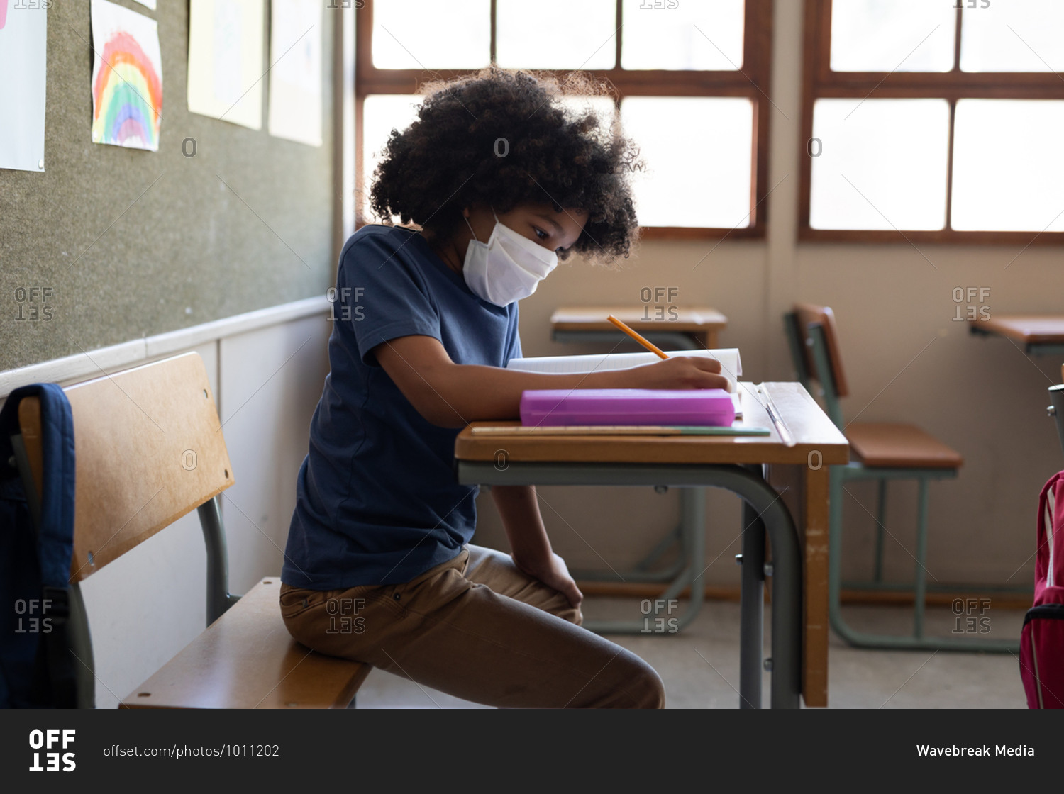 Mixed race boy sitting at desk wearing face mask in classroom. Primary education social distancing health safety during Covid19 Coronavirus pandemic.