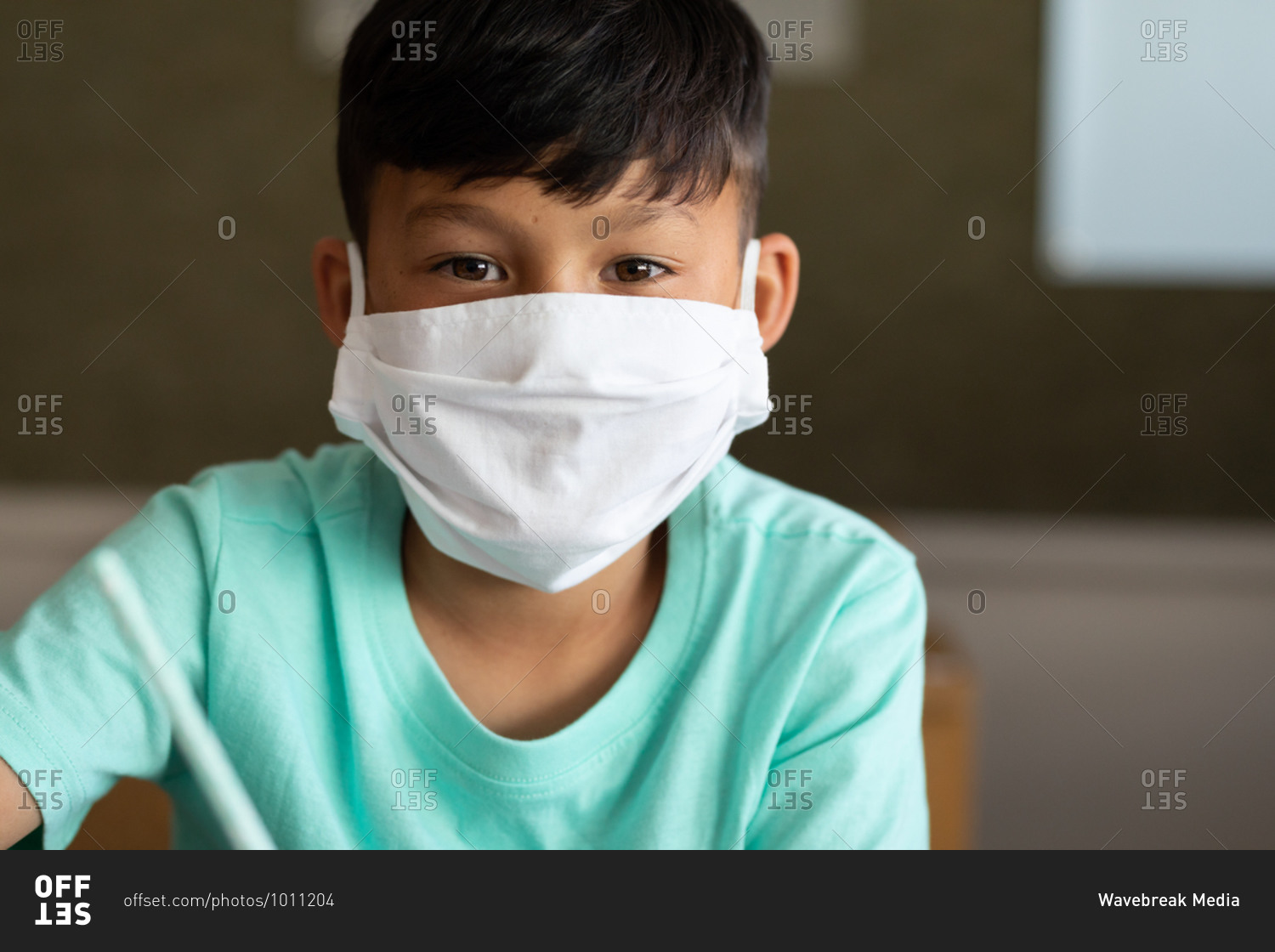 Portrait of an Asian boy sitting at desk wearing face mask in classroom. Primary education social distancing health safety during Covid19 Coronavirus pandemic.