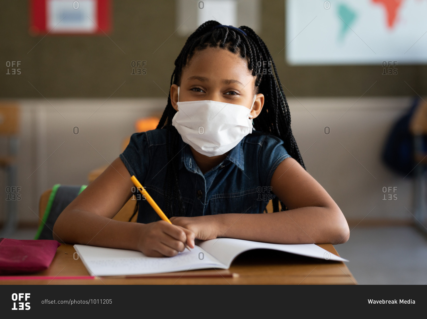 Portrait of a mixed race girl sitting at desk wearing face mask in classroom. Primary education social distancing health safety during Covid19 Coronavirus pandemic.