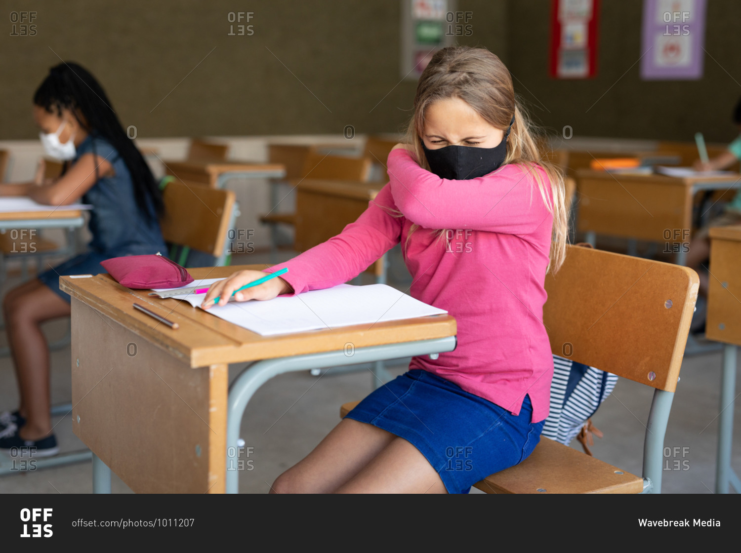 Caucasian girl sitting at desk wearing face mask in classroom, covering her face while sneezing. Primary education social distancing health safety during Covid19 Coronavirus pandemic.