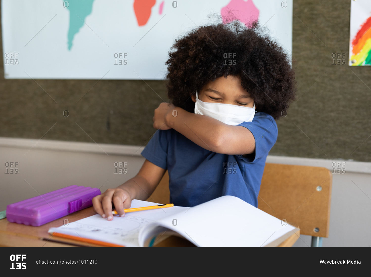 Mixed race boy sitting at desk wearing face mask in classroom, covering his face while sneezing. Primary education social distancing health safety during Covid19 Coronavirus pandemic.