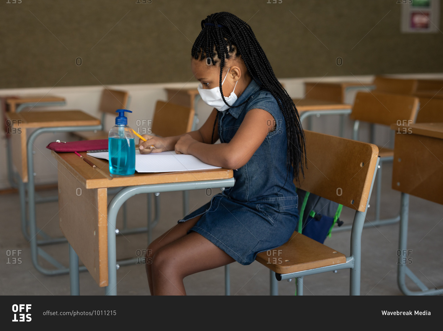 Mixed race girl wearing face mask while sitting on her desk at classroom with a sanitizer. Primary education social distancing health safety during Covid19 Coronavirus pandemic.