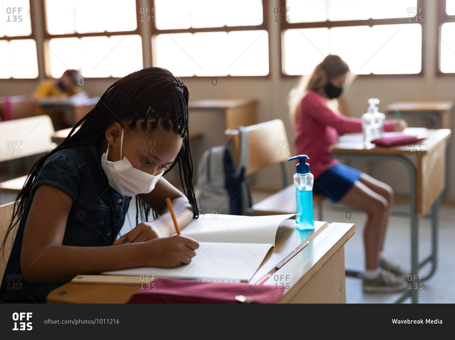 Mixed race girl wearing face mask while sitting on her desk at classroom with a sanitizer. Primary education social distancing health safety during Covid19 Coronavirus pandemic.