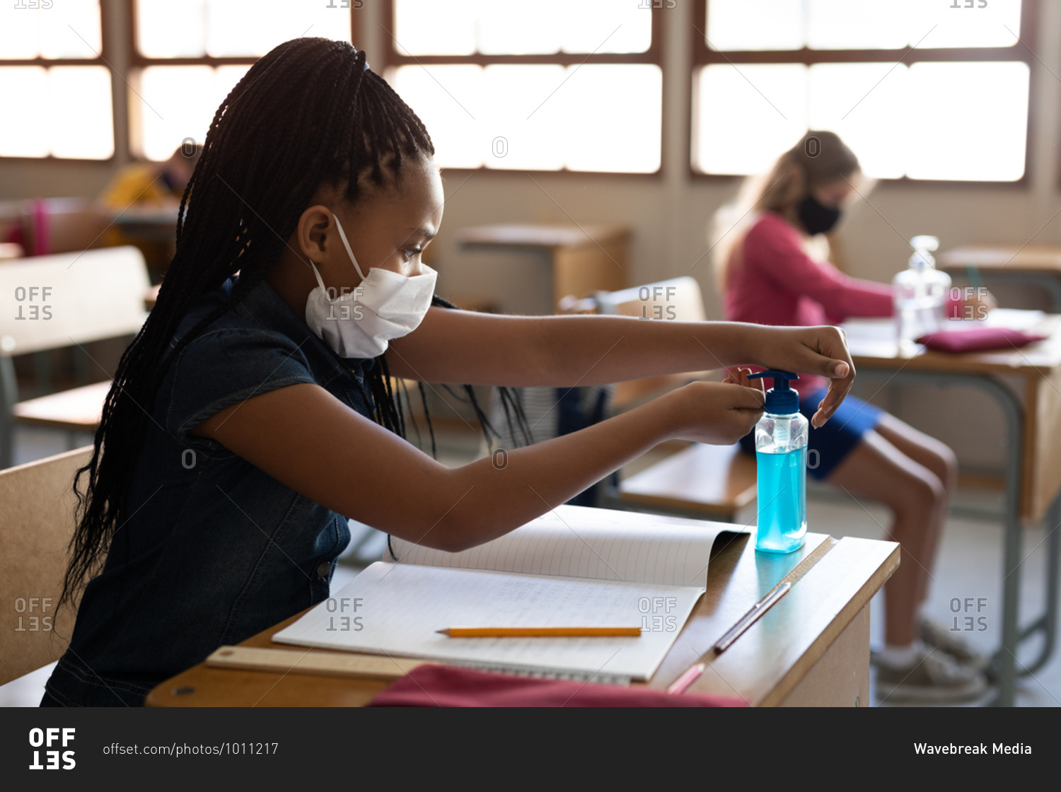 Mixed race girl wearing face mask, sanitizing her hands while sitting on her desk at classroom. Primary education social distancing health safety during Covid19 Coronavirus pandemic.