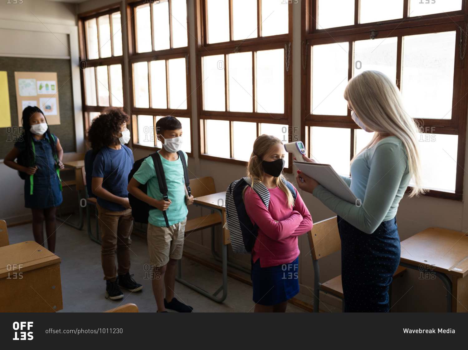 Caucasian female teacher wearing face mask measuring temperature of children in an elementary school. Primary education social distancing health safety during Covid19 Coronavirus pandemic.
