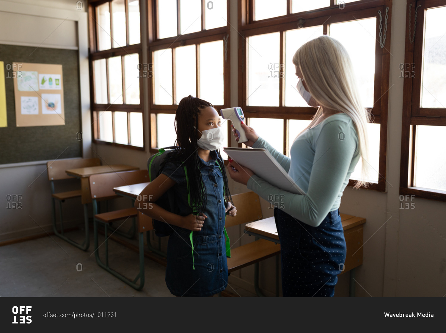 Caucasian female teacher wearing face mask measuring temperature of a girl in an elementary school. Primary education social distancing health safety during Covid19 Coronavirus pandemic.