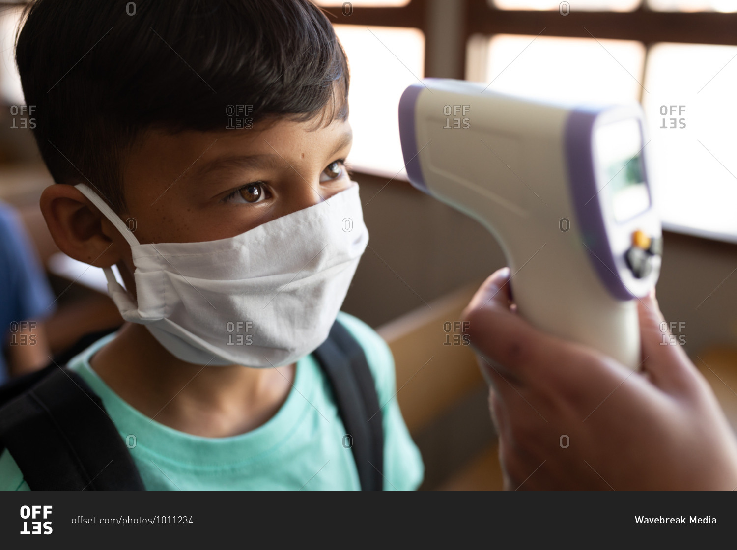 Mixed race boy wearing face mask getting his temperature measured in an elementary school. Primary education social distancing health safety during Covid19 Coronavirus pandemic.