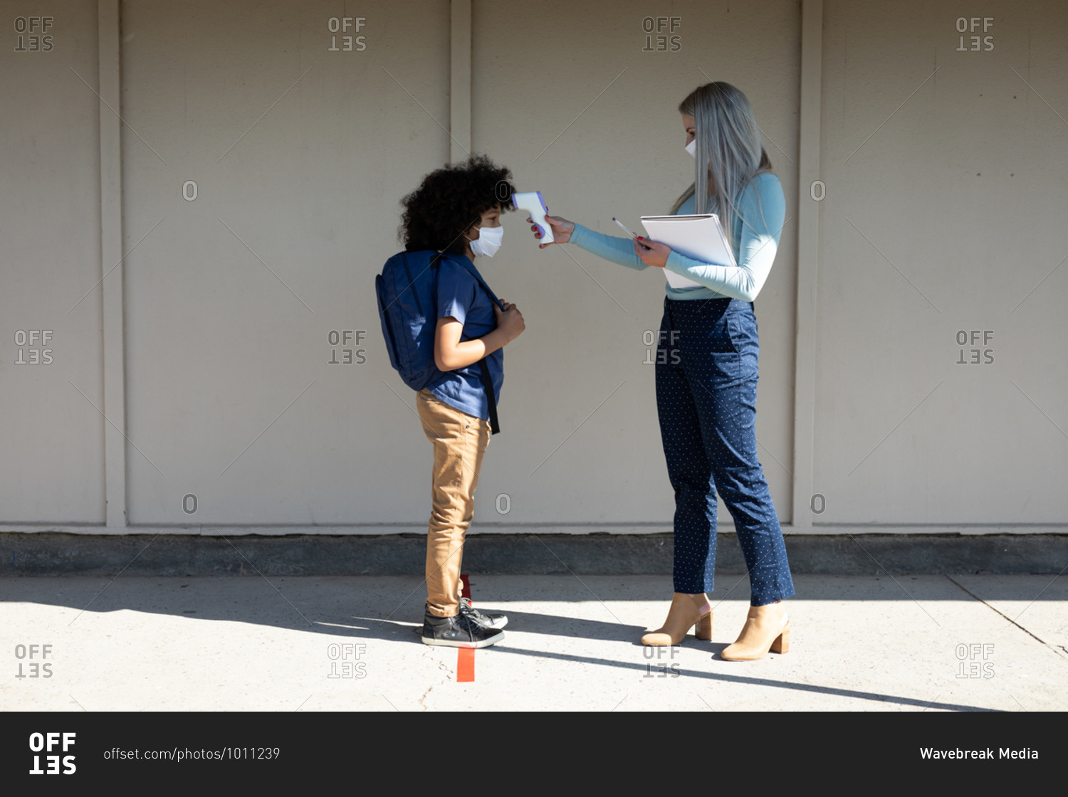 Caucasian female teacher wearing face mask measuring temperature of a boy in an elementary school. Primary education social distancing health safety during Covid19 Coronavirus pandemic.