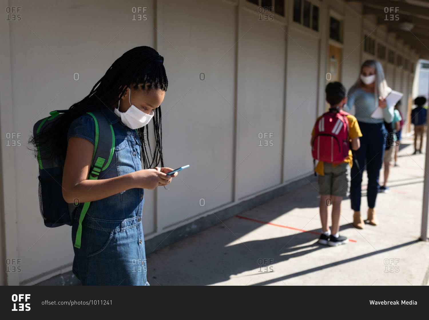 Mixed race girl wearing face mask using smartphone and teacher measuring temperature in elementary school. Primary education social distancing health safety during Covid19 Coronavirus pandemic.