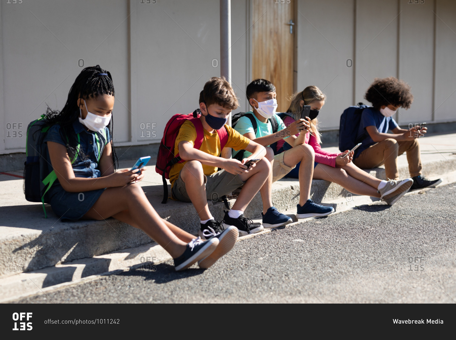 Multi ethnic group of elementary school kids wearing face masks using smartphones while sitting together. Primary education social distancing health safety during Covid19 Coronavirus pandemic.
