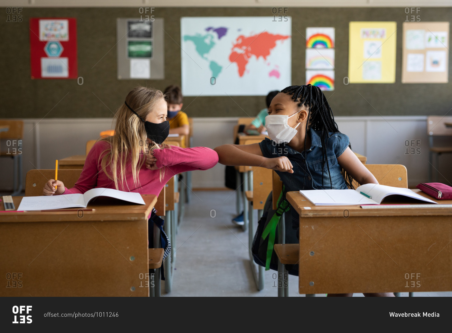 Two multi ethnic girls wearing face masks greeting each other by touching elbows in the classroom. Primary education social distancing health safety during Covid19 Coronavirus pandemic.
