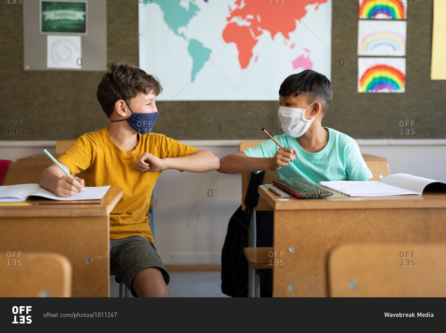 Two multi ethnic boys wearing face masks greeting each other by touching elbows in the classroom. Primary education social distancing health safety during Covid19 Coronavirus pandemic.