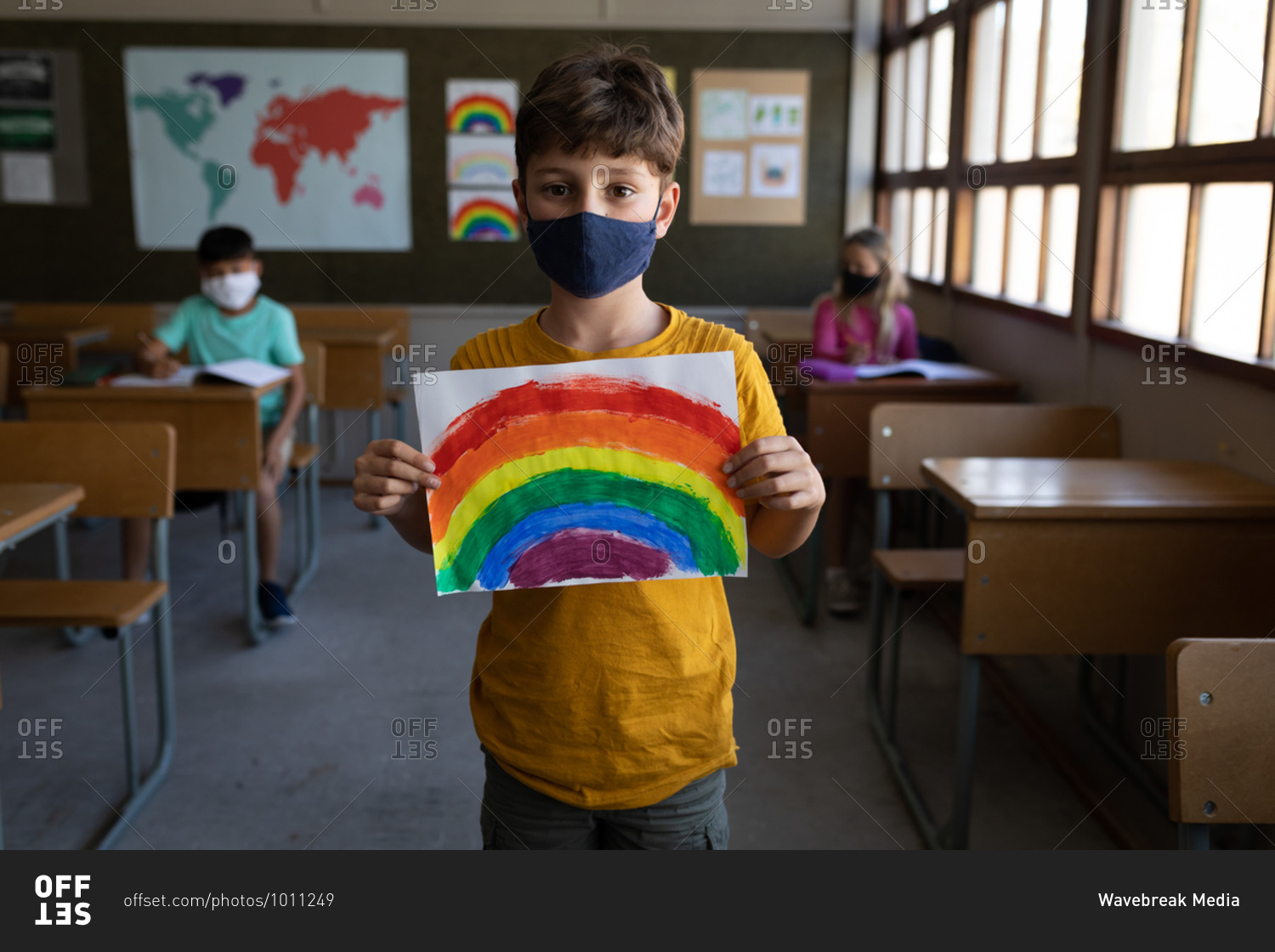 Portrait of a Caucasian boy wearing face mask holding a rainbow drawing in the classroom. Primary education social distancing health safety during Covid19 Coronavirus pandemic.