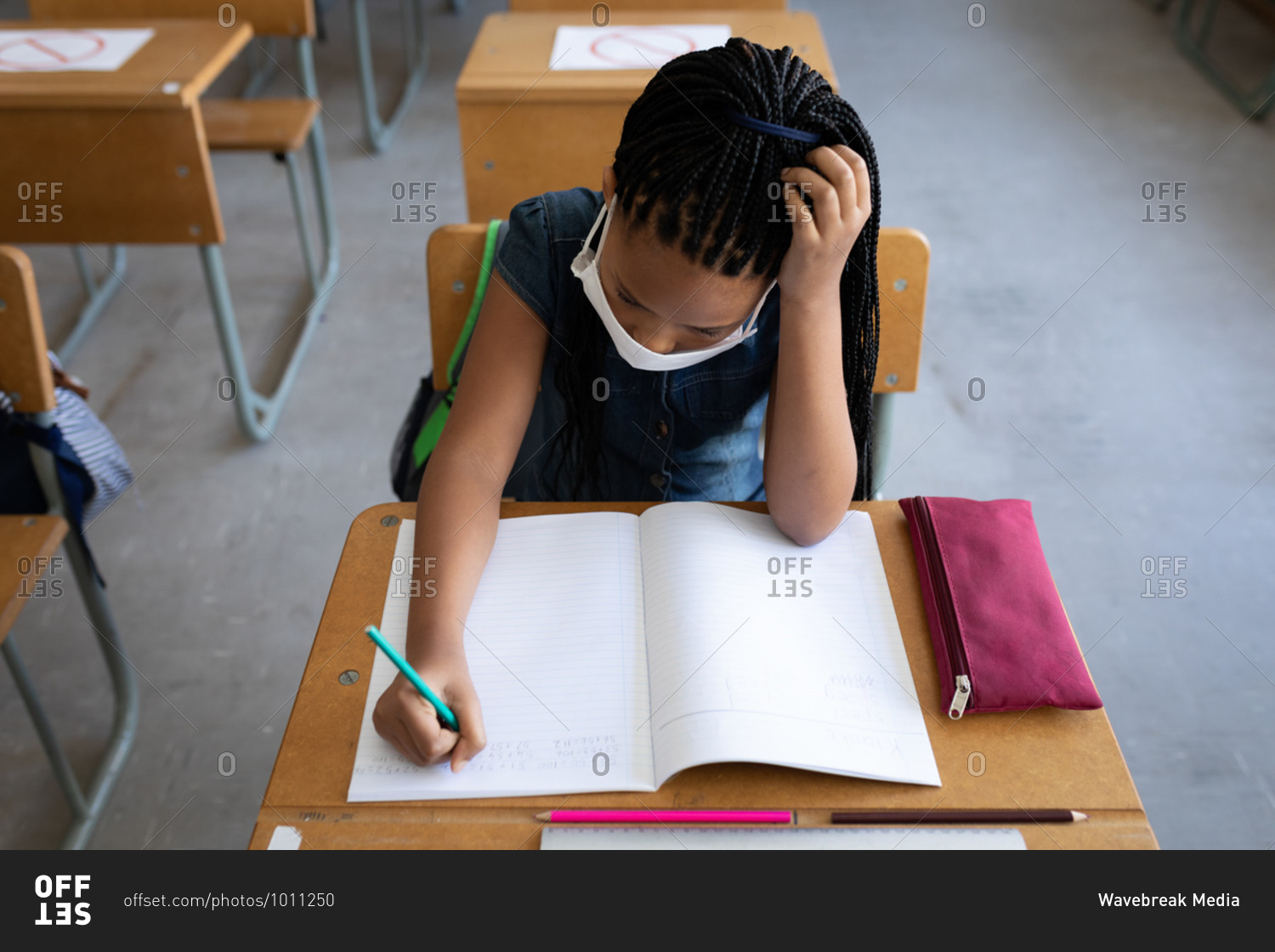 Overhead view of a mixed race girl wearing face mask writing while sitting on her desk in the classroom. Primary education social distancing health safety during Covid19 Coronavirus pandemic.