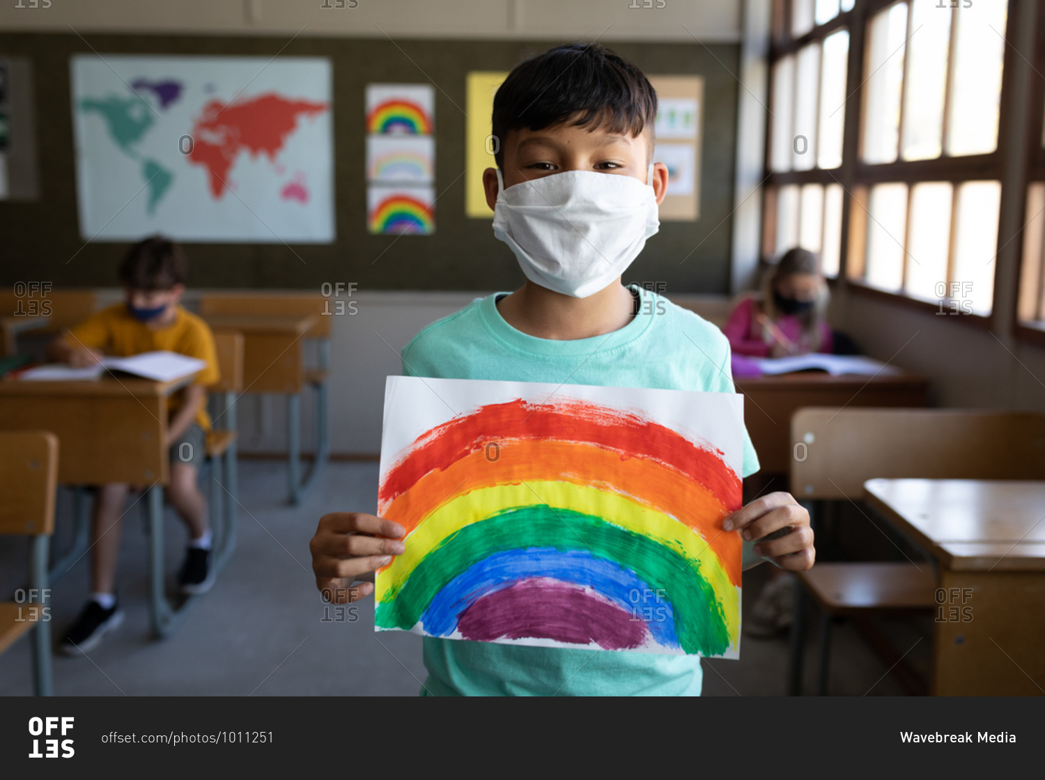 Portrait of a mixed race boy wearing face mask holding a rainbow drawing in the classroom. Primary education social distancing health safety during Covid19 Coronavirus pandemic.