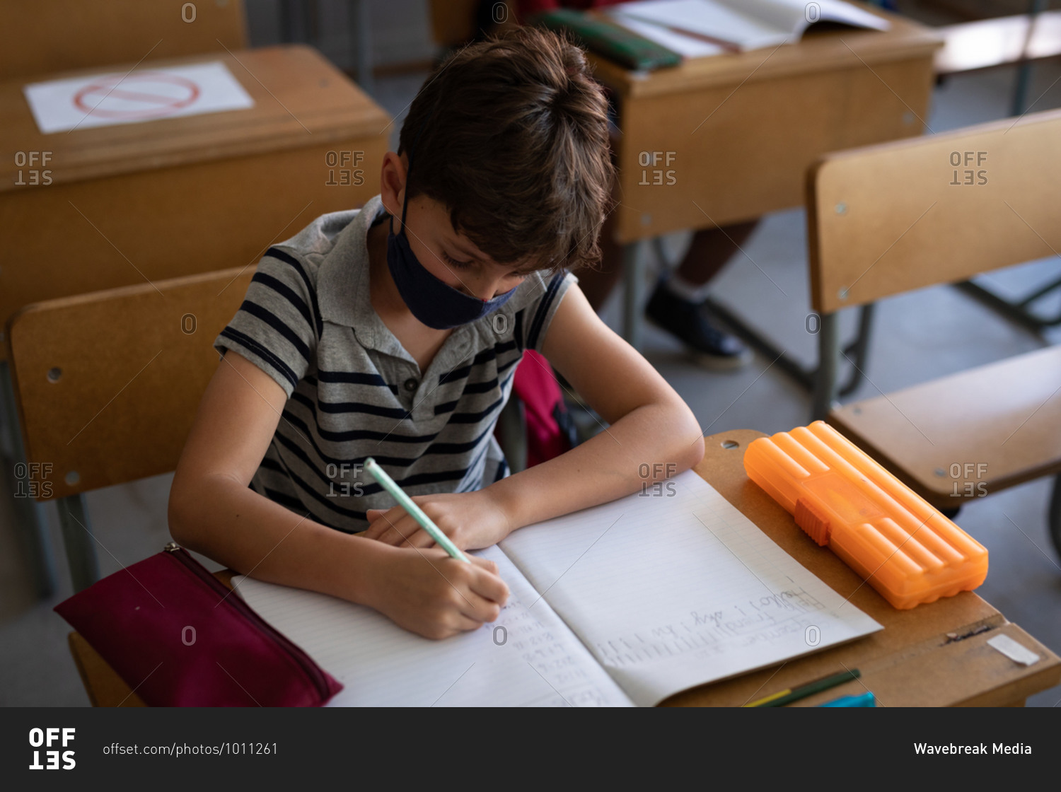 Caucasian boy wearing face mask, sitting on his desk during the lesson. Primary education social distancing health safety during Covid19 Coronavirus pandemic.