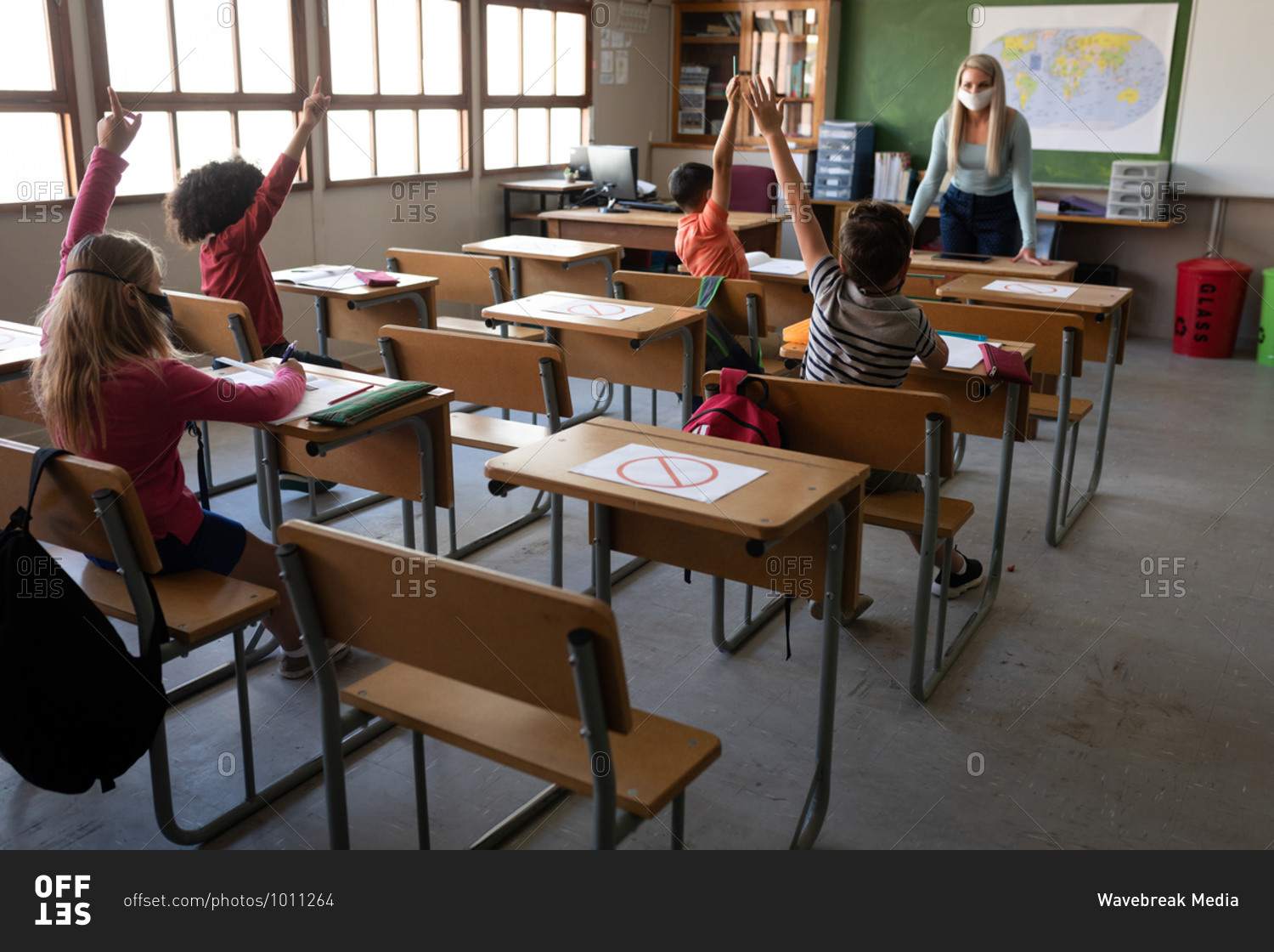 Group of multi ethnic kids sitting on their desk during the lesson with a female teacher wearing a face mask. Primary education social distancing health safety during Covid19 Coronavirus pandemic.