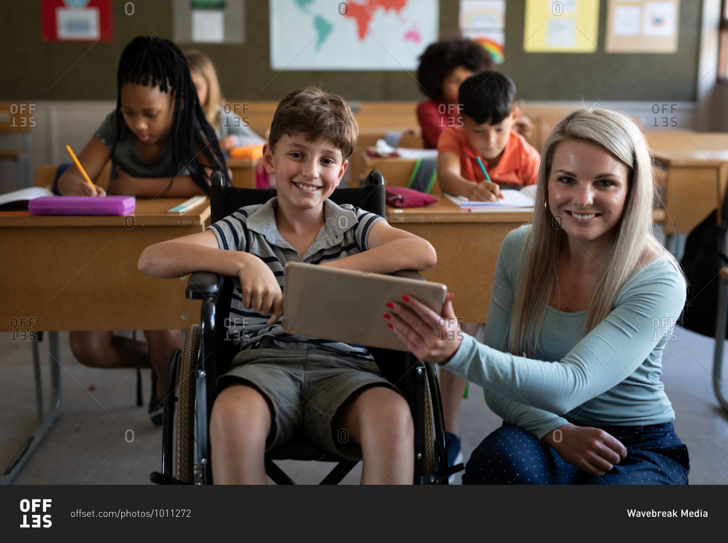 Portrait of disable Caucasian boy sitting in his wheelchair and his female teacher using tablet in the classroom. Primary education social distancing health safety during Covid19 Coronavirus pandemic.