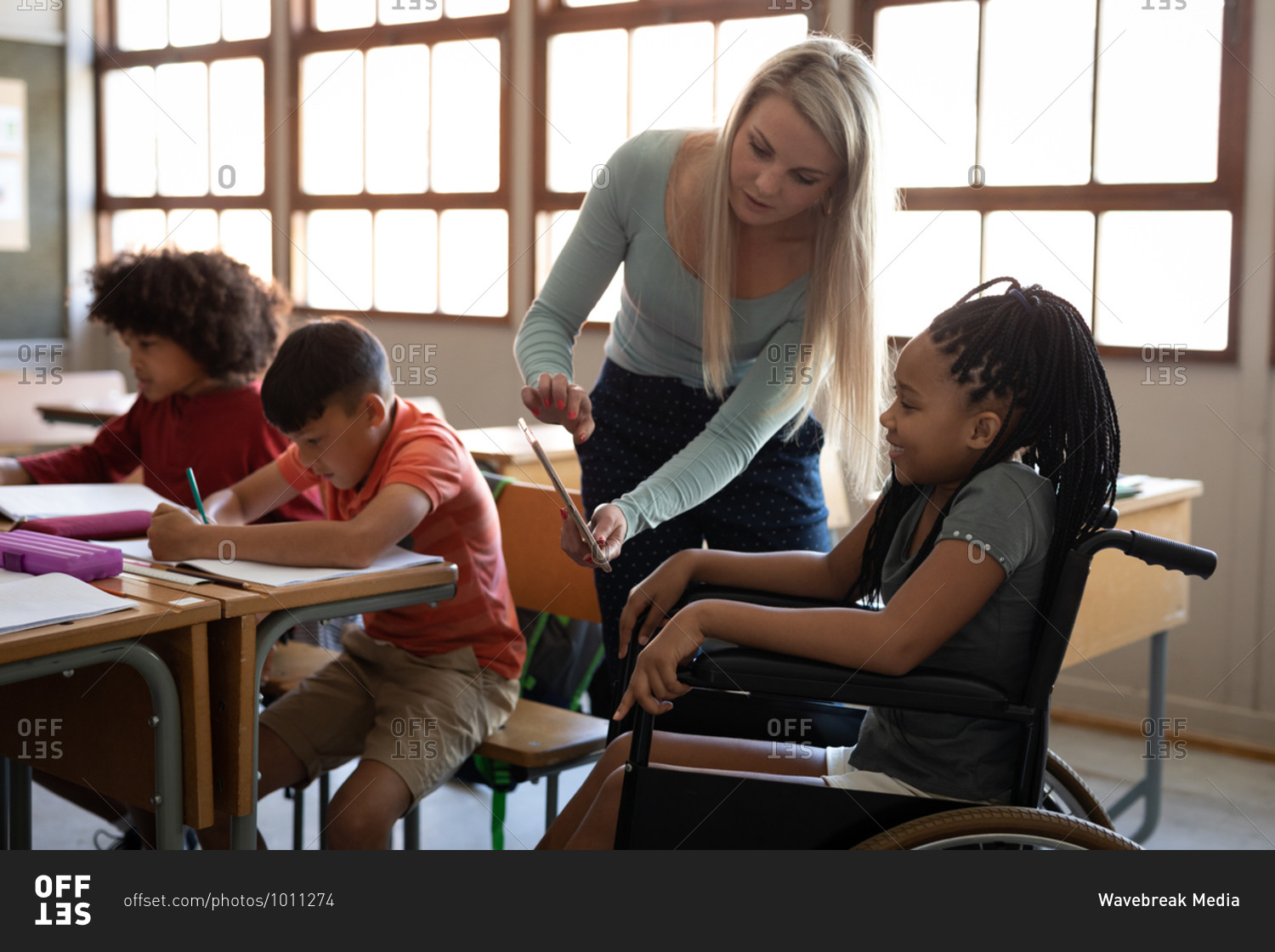 Disable mixed race girl sitting in her wheelchair and her female teacher using tablet in the classroom. Primary education social distancing health safety during Covid19 Coronavirus pandemic.
