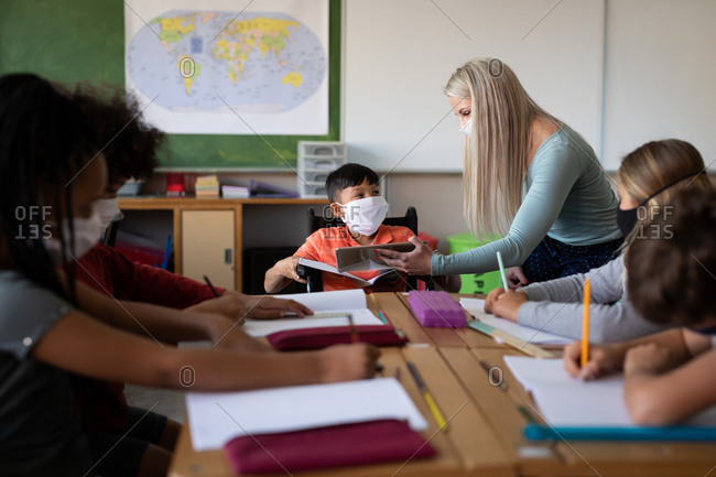 Caucasian female teacher wearing face mask with digital tablet teaching group of multi ethnic kids. Primary education social distancing health safety during Covid19 Coronavirus pandemic.