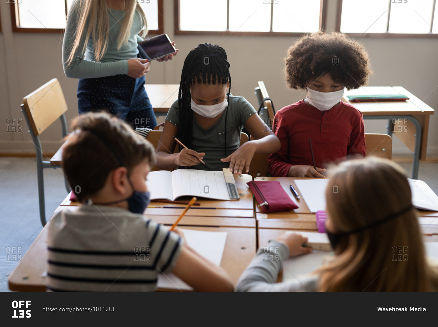 Caucasian female teacher wearing face mask teaching group of multi ethnic kids. Primary education social distancing health safety during Covid19 Coronavirus pandemic.