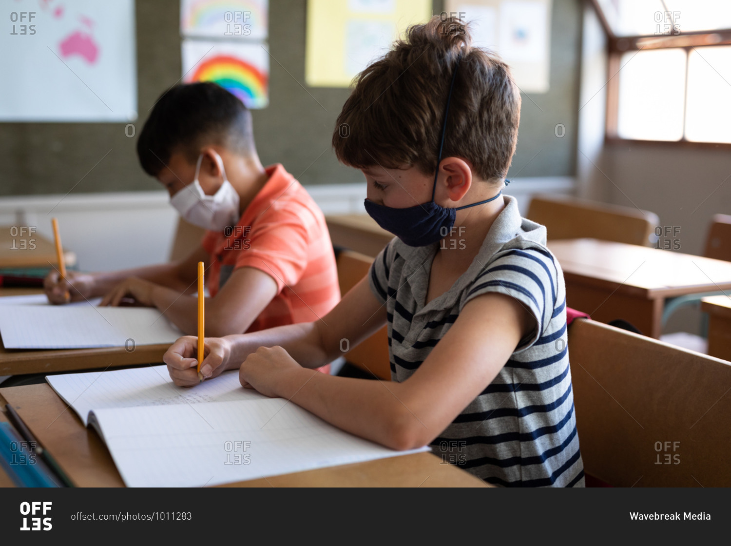 Two multi ethnic boys sitting at desks wearing face masks in classroom. Primary education social distancing health safety during Covid19 Coronavirus pandemic.