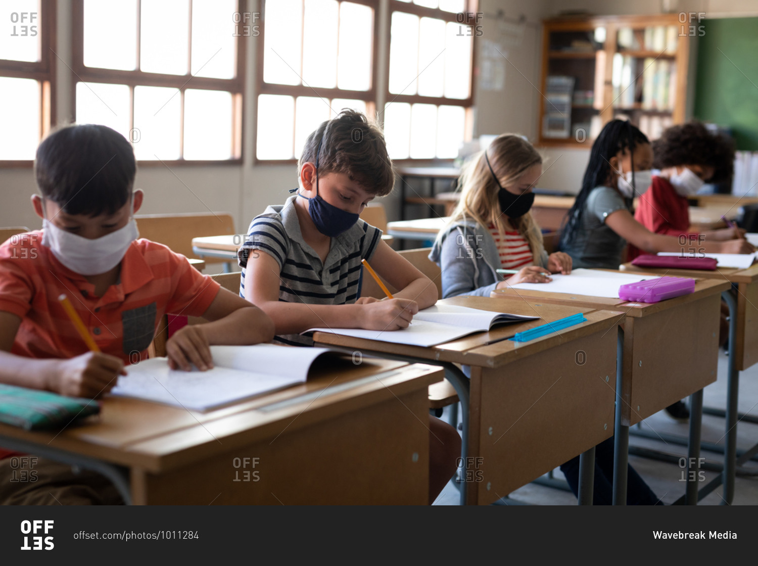 Multi ethnic group of elementary school children sitting at desks wearing face masks in classroom. Primary education social distancing health safety during Covid19 Coronavirus pandemic.