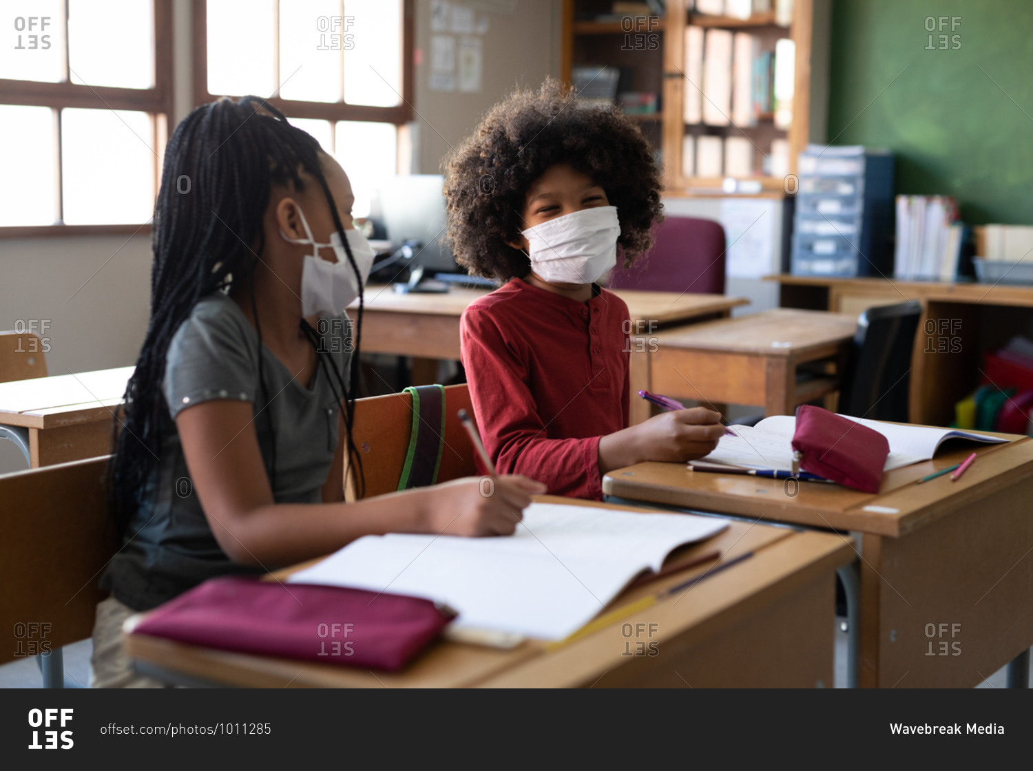 Multi ethnic boy and girl sitting at desks wearing face masks in classroom. Primary education social distancing health safety during Covid19 Coronavirus pandemic.