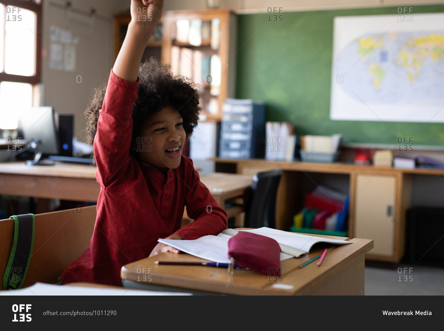Mixed race boy raising his hand while sitting on his desk at school. Primary education social distancing health safety during Covid19 Coronavirus pandemic.