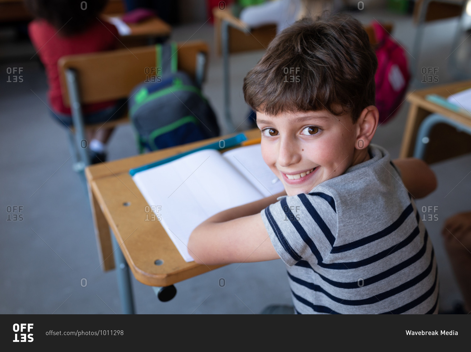 Portrait of a Caucasian boy smiling while sitting on his desk at school. Primary education social distancing health safety during Covid19 Coronavirus pandemic.