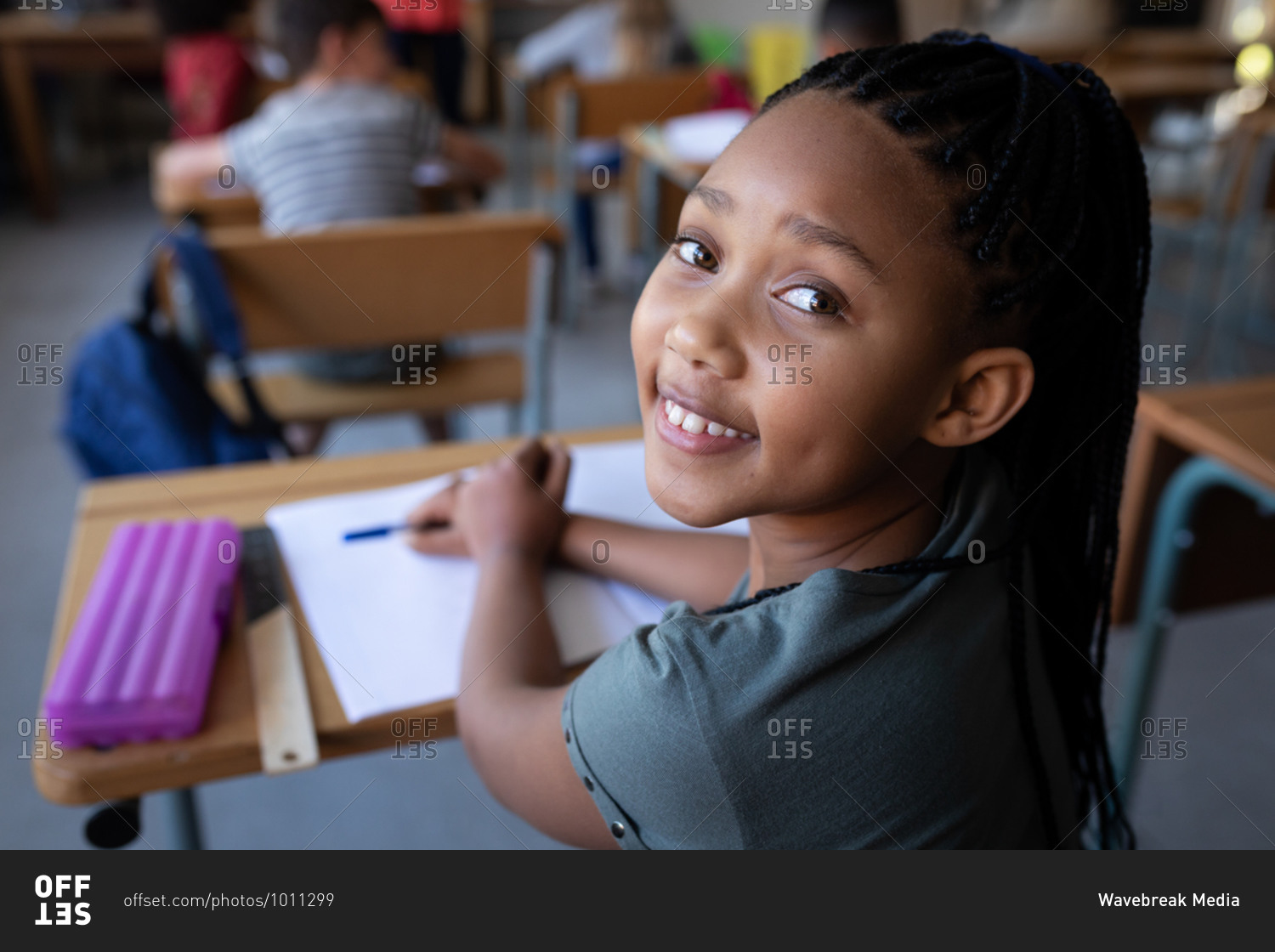 Portrait of a mixed race girl smiling while sitting on her desk at school. Primary education social distancing health safety during Covid19 Coronavirus pandemic.