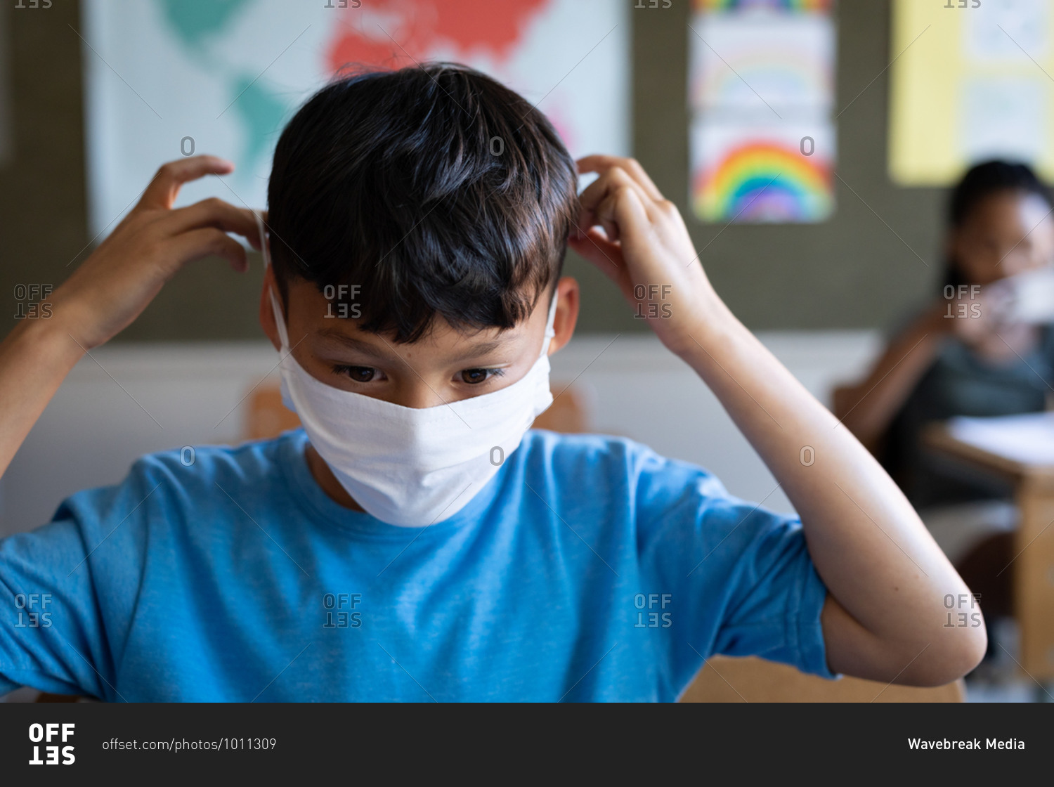 Mixed race boy wearing face mask while sitting on his desk at school. Primary education social distancing health safety during Covid19 Coronavirus pandemic.