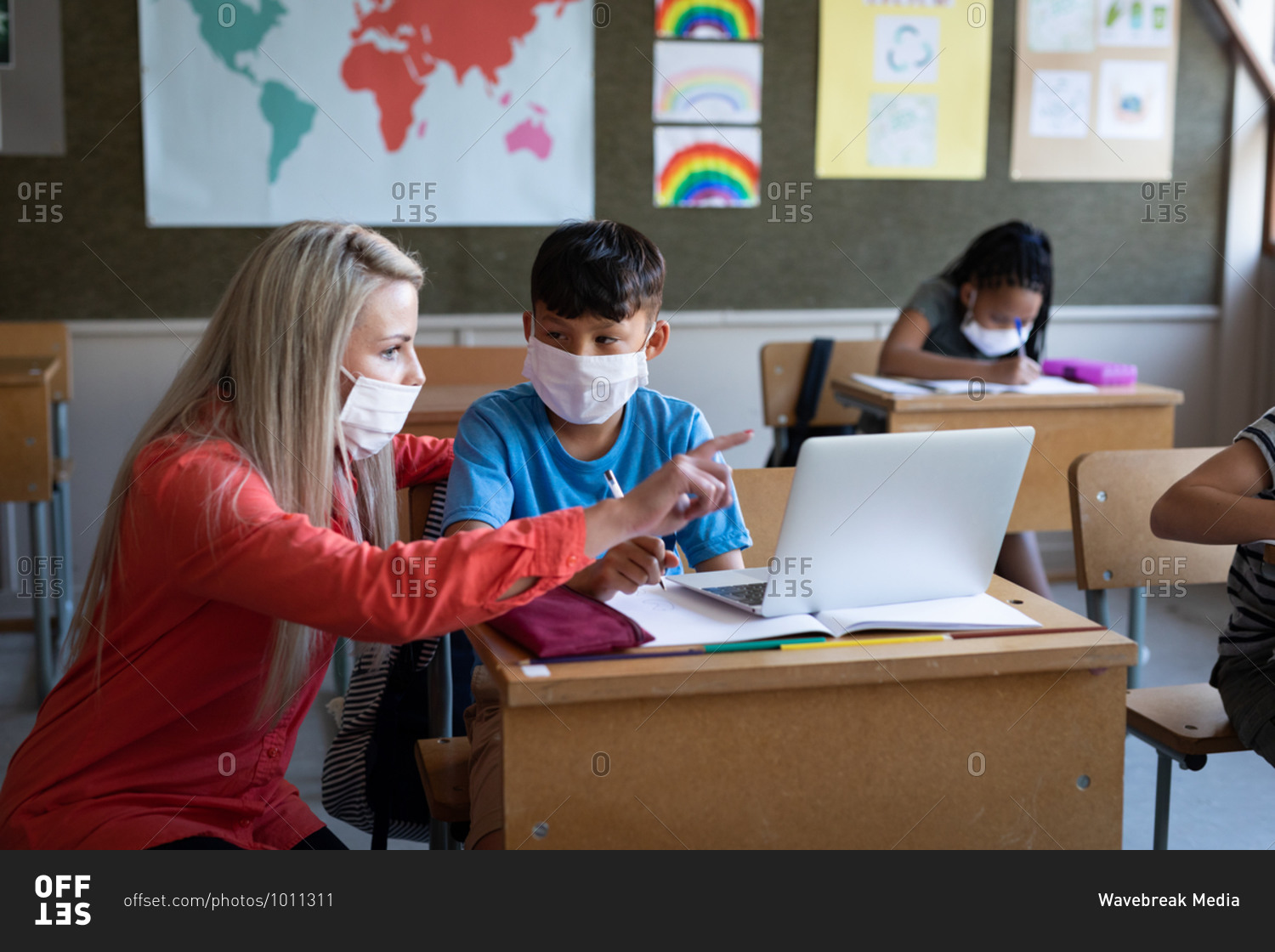 Female Caucasian teacher and mixed race boy wearing face masks using laptop in class at school. Primary education social distancing health safety during Covid19 Coronavirus pandemic.
