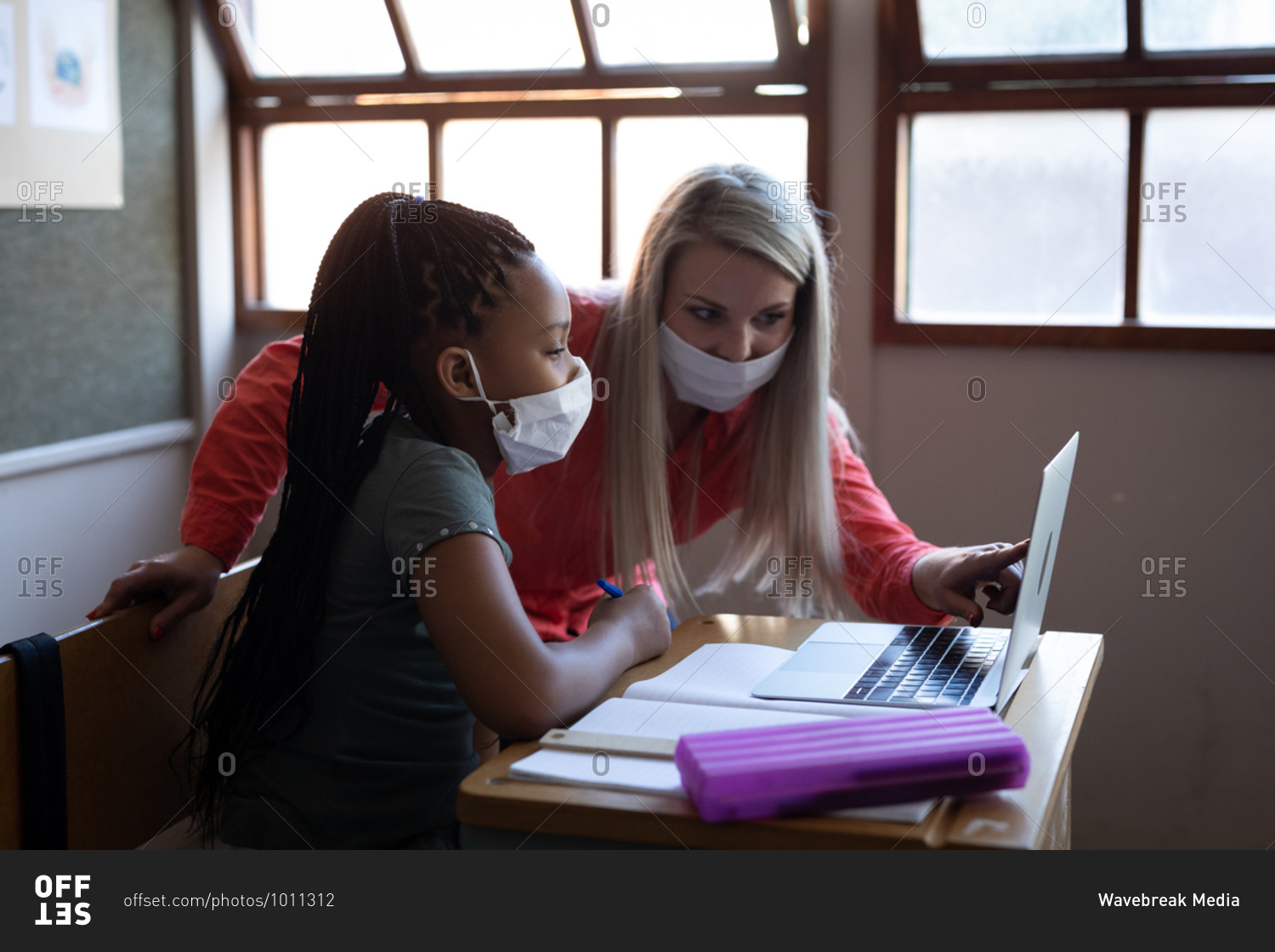 Female Caucasian teacher and mixed race girl wearing face masks using laptop in class at school. Primary education social distancing health safety during Covid19 Coronavirus pandemic.