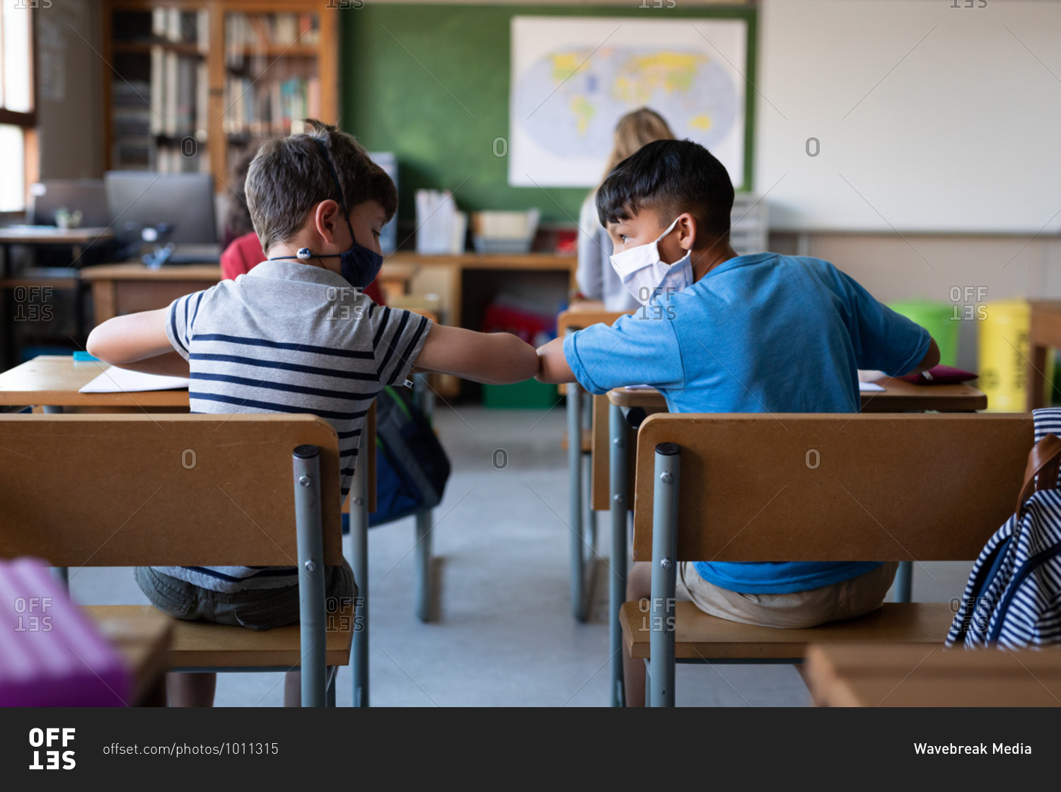 Two multi ethnic boys wearing face masks greeting each other by touching elbows at school. Primary education social distancing health safety during Covid19 Coronavirus pandemic.