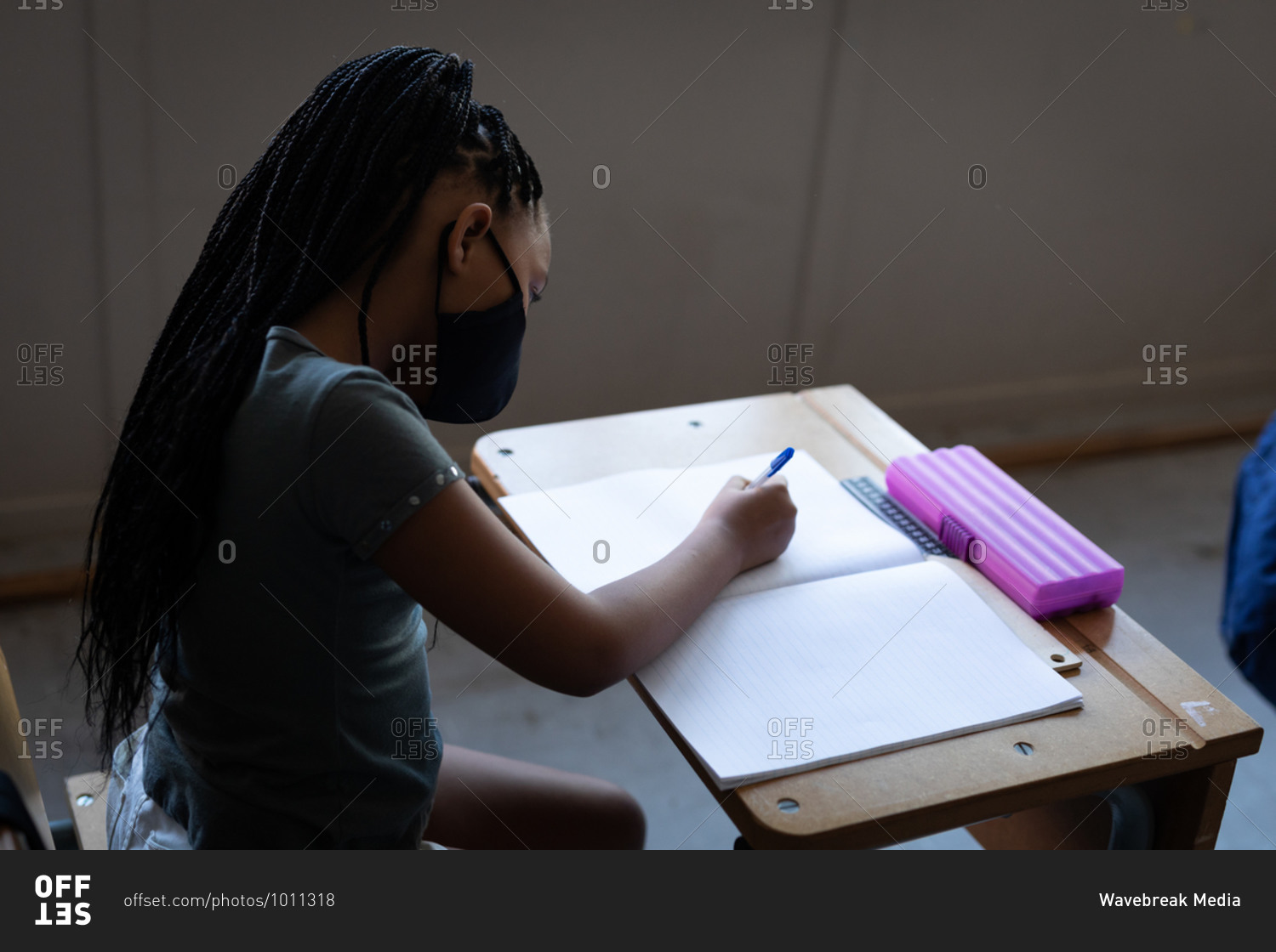 Mixed race girl wearing face mask writing while sitting on her desk at school. Primary education social distancing health safety during Covid19 Coronavirus pandemic.