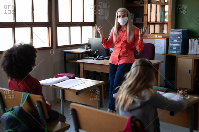 Female Caucasian teacher wearing face mask teaching in class at school. Primary education social distancing health safety during Covid19 Coronavirus pandemic.