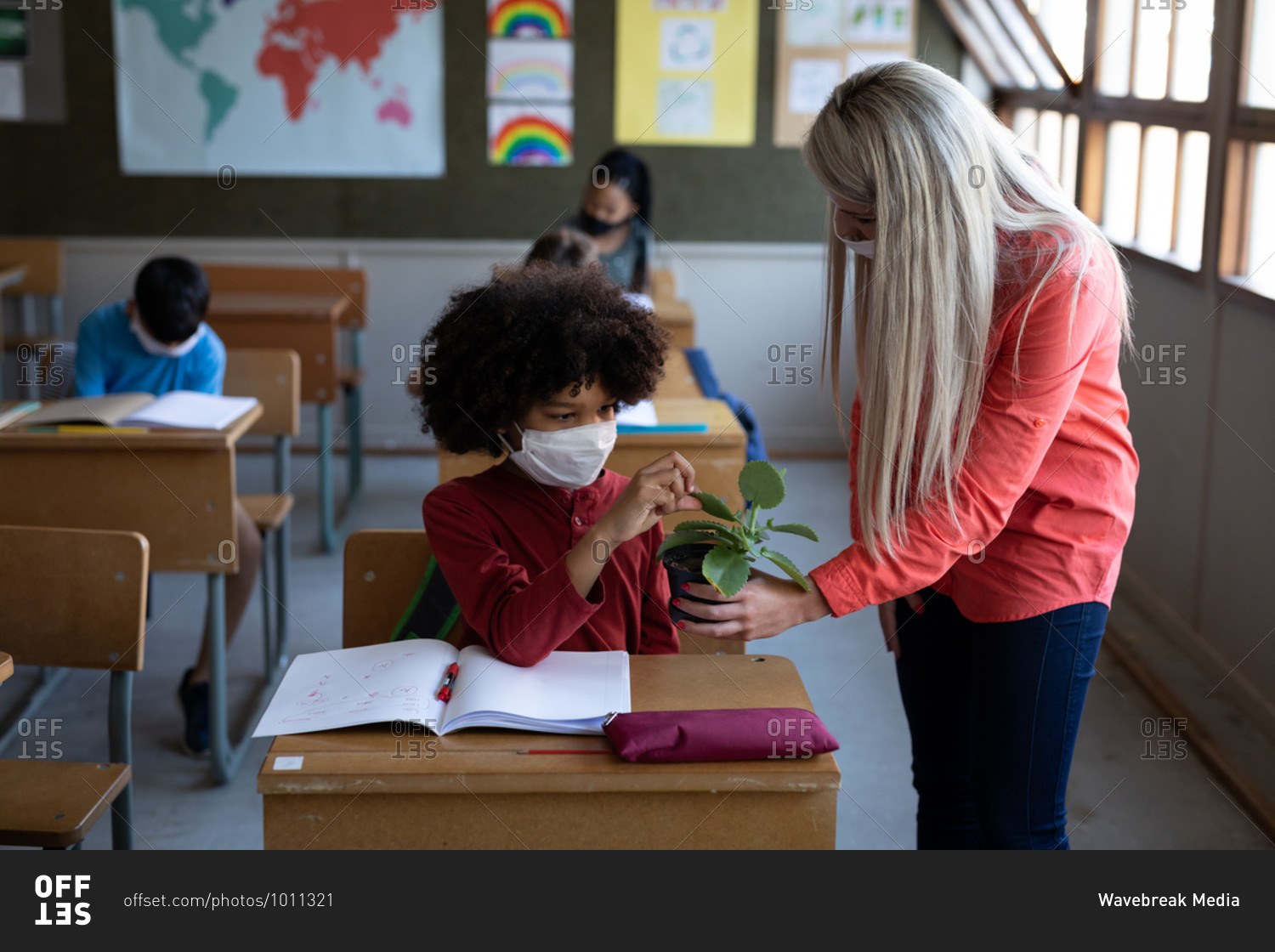 Female Caucasian teacher wearing face mask showing a plant pot to a mixed race boy in school. Primary education social distancing health safety during Covid19 Coronavirus pandemic.